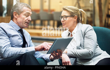 Reifen Geschäftsmann und Geschäftsfrau, sitzen auf Stühlen in einem Büro Lobby arbeiten zusammen und diskutieren über ein digitales Tablet Stockfoto