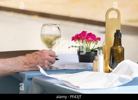 Eine männliche Hand mit einem Glas Weißwein auf einem Tisch eines Restaurants im Freien platziert. Stockfoto