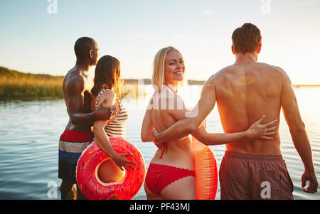 Lächelnde Frau im Bikini über die Schulter schauen beim Stehen mit ihrem Freund und einem anderen Paar in einem See den Sonnenuntergang zu beobachten. Stockfoto