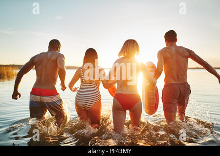 Rearview eine vielfältige Gruppe von jungen Freunden in Badebekleidung in einen See zusammen laufen bei Sonnenuntergang Spritzwasser. Stockfoto