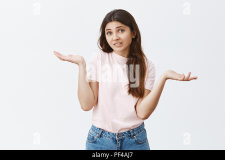 Innen- schuss von verwirrt und unsicher, attraktiven europäischen weiblichen Brünette in stilvollem Rosa t-shirt zuckte mit den Händen zur Seite ausbreiten und schuldig, Lächeln, ratlos und unsicher über grauer Hintergrund Stockfoto