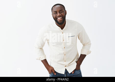 Charmant lustig African American reifen männlichen versuchen, Witze, cute woman beeindrucken, schmunzelt und breit grinsend, kippen in Richtung Kamera, Hände in den Taschen, die wenig Scheu über graue Wand Stockfoto