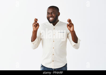 Heute Glück ist auf meiner Seite. Nervös hoffnungsvoll lustig African American in weiß casual Shirt holding Daumen und Aufeinanderpressen der Zähne beim Wunsch nach Wunsch wahr werden über grauer Hintergrund Stockfoto