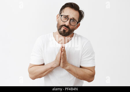 Bitte ich ihre Hilfe jetzt brauche. Portrait von umgekippt und düsteren gut aussehender Mann in einer verzweifelten Situation, Holding hands in beten und runzelte die Stirn und bitten, betteln um Vergebung über die graue Wand Stockfoto