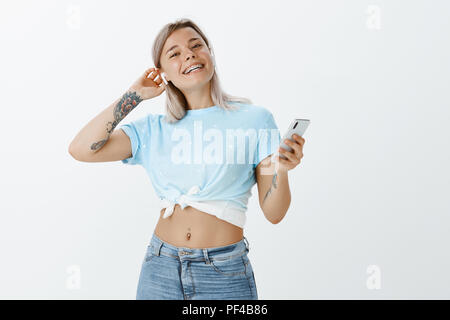 Schauen Sie, was ich für mich gekauft. Portrait von stilvollen glücklich Frau mit tätowierten Arm in stilvoller Kleidung, Smartphone, berühren Haar und Drahtlose Ohrhörer, breit grinsend Stockfoto