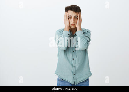 Portrait von depressiv unglücklich Kerl in Trendy Shirt, die Hände auf den Kopf und starrte mit Gleichgültigkeit an Kamera, leiden Schlaflosigkeit oder Stress, Müdigkeit und über grauer Hintergrund zugeführt Stockfoto