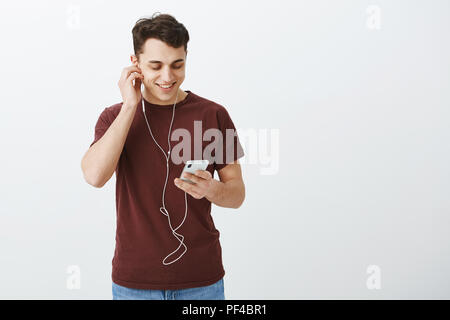 Innen- schuss von zufriedenen Sorglos gut aussehender Kerl in casual rotes T-Shirt, Ohrhörer und breit grinsend am Bildschirm des Smartphones während Pop Musik und genießen Sie hochwertigen Sound Stockfoto