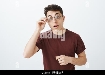 Studio shot von Smart neugierig stattlicher Mann in runde Gläser und rotes T-Shirt, Anhebung der Augenbrauen mit Interesse und Holding rim Brille sehen interessanter Fall, wollen zu lösen Rätsel oder Zuordnung tun Stockfoto