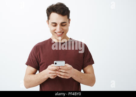 Taille-up Shot der positiven freundlich aussehenden Mann im roten T-Shirt, Smartphone und freudig lächelnd an Bildschirm beim Senden lustiges Bild, Freundin, die gemeinsame Nutzung von Memen und Lachen Stockfoto