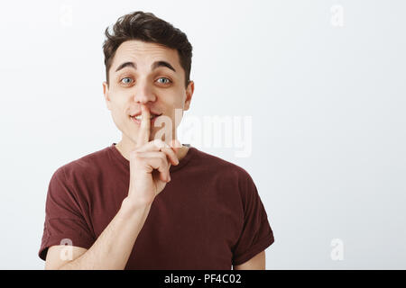 Portrait von charmanten verspielten männliche Kursteilnehmer im Casual Trendy T-Shirt, Hebezeuge eyebrown mit glücklichen freundlich Ausdruck, sagen Shh und zeigt Psst mit Zeigefinger über Mund, neugierig lächelnd Stockfoto