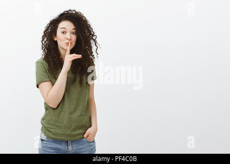 Unser kleines Geheimnis bleiben. Portrait von verspielten faszinierende attraktives Mädchen mit lockigem Haar, sagen Shh und psst Geste mit dem Zeigefinger über Mund, sagt Klatsch heimlich über grauer Hintergrund Stockfoto