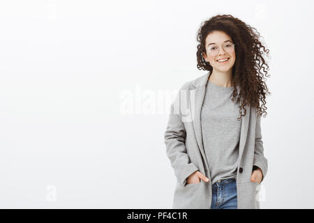 Mädchen warten, während Freund unter Bild. Modisch gut aussehende Frau in Gläser und stilvolle Mantel, die Hände in den Taschen, breit grinsend, Vertrauen und die freundliche Haltung Stockfoto