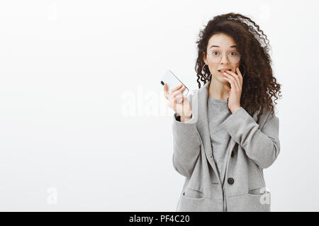 Portrait von peinlich cute Curly blonde Studentin im stilvollen grauen Mantel und Gläser, Smartphone, Anheben der Augenbrauen und beißen Finger, machen Fehler und Gefühl über graue Wand unbeholfen Stockfoto