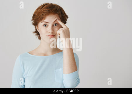 Ich bin nicht überrascht, es war offensichtlich. Studio shot von attraktiven Mädchen mit Ingwer Haar Anheben der Augenbrauen mit Zeigefinger, grinsend und Gefühl unbeholfen und unbeeindruckt, stehend gegen grauen Hintergrund. Stockfoto