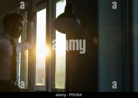Seitenansicht eines Geschäftsmannes tragen weiße Hemd und schwarzer Krawatte, während durch das Fenster bei Sonnenuntergang. Stockfoto