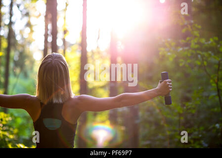 Junge blonde Frau, die die Arbeit mit Hanteln in schöner Natur mit dem Rücken zur Kamera und die Arme weit verbreitet. Konzeptionelle der Übung und Körper Stockfoto