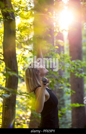 Gerne blonde junge Frau tun Pilates Übung in einem Wald bei Sonnenaufgang mit Hanteln, Stretching ein Arm hoch in die Luft. Stockfoto