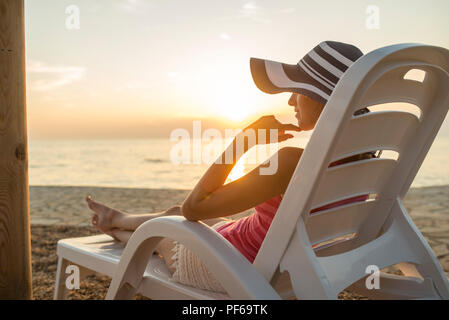 Junge Frau mit sonnenhut Sitzen auf einem Liegestuhl mit Blick auf das Meer bei Sonnenuntergang. Stockfoto