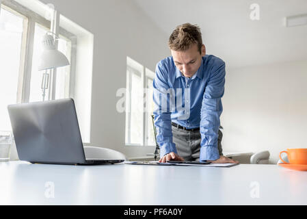 Stilvolle Junge Unternehmer stehend lehnte sich auf seinen Schreibtisch lesen um einen Tablet-PC mit einem ernsten Ausdruck, Vorderansicht. Stockfoto