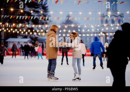 Junges Paar in Liebe kaukasischen Mann mit blondem Haar mit langen Haaren und Bart und schöne Frau Spaß haben, aktive Datum Eislaufen Auf dem Eis Arena in Stockfoto