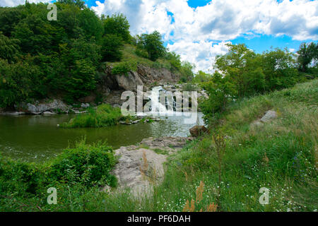 Der Wasserfall auf dem Fluss fließt durch und über die Felsen bedeckt mit Flechten und Moos vor dem hintergrund der grünen Vegetation und einem blauen Himmel. Stockfoto