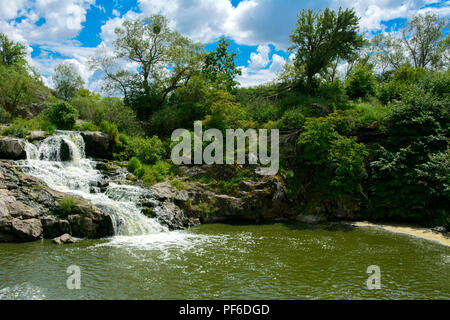 Der Wasserfall auf dem Fluss fließt durch und über die Felsen bedeckt mit Flechten und Moos vor dem hintergrund der grünen Vegetation und einem blauen Himmel. Stockfoto