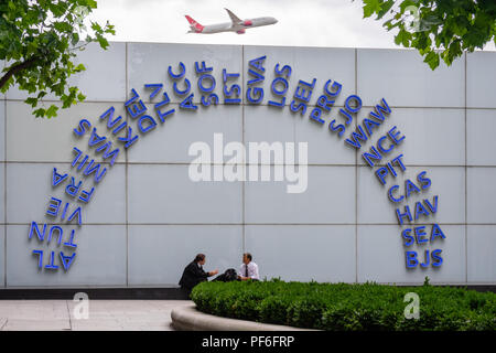 Blau beleuchtete IATA-Codes vor dem Terminal 5 des Flughafens London Heathrow, ein Virgin Atlantic Flugzeug, das im Hintergrund abhebt Stockfoto