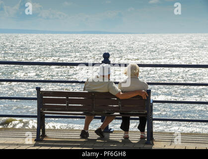 Älterer Mann und Frau sitzt auf einer Holzbank an der Promenade in Porthcawl, Wales, mit Blick auf den Kanal von Bristol Stockfoto