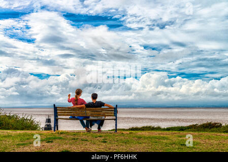 Ein paar sitzen unter einem bewölkten Himmel auf Battery Point, Portishead in der Nähe von Bristol, England, Großbritannien mit Blick auf den Severn Estuary mit Wales darüber hinaus. Stockfoto