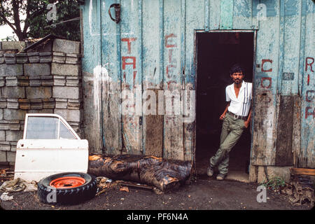 Managua, Nicaragua, Juli 1986; ein Junge rollt ein Auto, Rad, das durch die Run repariert wurde Mechanik Werkstatt hinter sich. Stockfoto