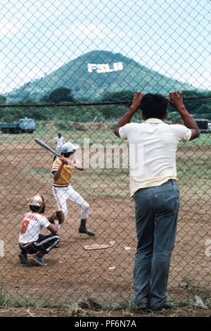 Managua, Nicaragua, Juni 1986; Leute Baseball zu spielen. Die Beschilderung der Sandinistischen FSLN ist in großen Buchstaben sichtbar auf dem gegenüberliegenden Hang. Stockfoto