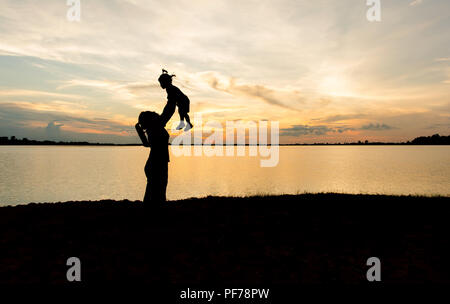 Silhouette von Mutter und Tochter anheben Kind in der Luft über einen malerischen Sonnenuntergang Himmel am Flußufer. Entspannung und Glück Stockfoto