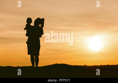 Silhouette von Mutter und Tochter anheben Kind in der Luft über einen malerischen Sonnenuntergang Himmel am Flußufer. Entspannung und Glück Stockfoto