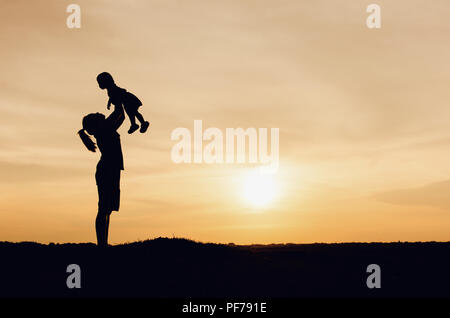 Silhouette von Mutter und Tochter anheben Kind in der Luft über einen malerischen Sonnenuntergang Himmel am Flußufer. Entspannung und Glück Stockfoto