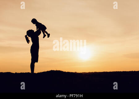 Silhouette von Mutter und Tochter anheben Kind in der Luft über einen malerischen Sonnenuntergang Himmel am Flußufer. Entspannung und Glück Stockfoto