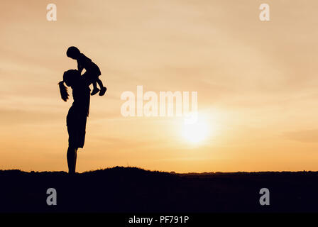 Silhouette von Mutter und Tochter anheben Kind in der Luft über einen malerischen Sonnenuntergang Himmel am Flußufer. Entspannung und Glück Stockfoto