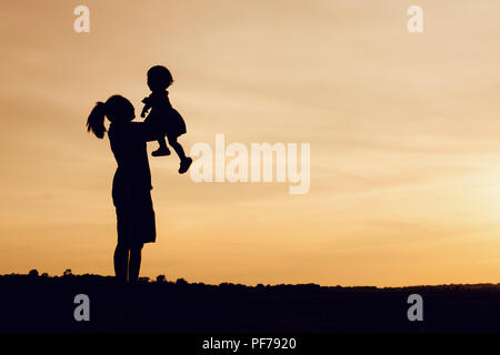 Silhouette von Mutter und Tochter anheben Kind in der Luft über einen malerischen Sonnenuntergang Himmel am Flußufer. Entspannung und Glück Stockfoto