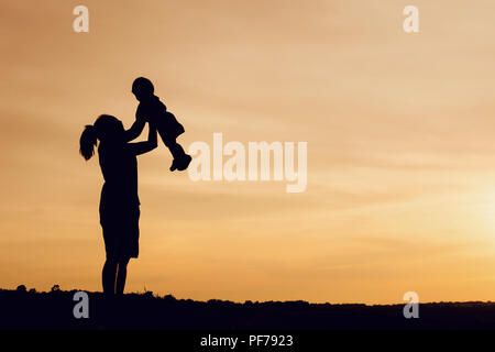 Silhouette von Mutter und Tochter anheben Kind in der Luft über einen malerischen Sonnenuntergang Himmel am Flußufer. Entspannung und Glück Stockfoto