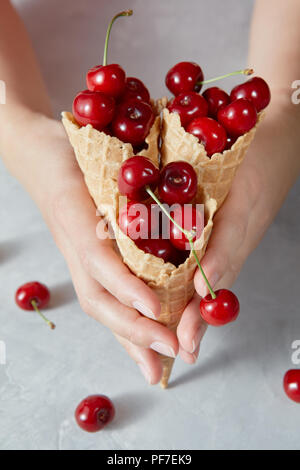 Weibliche halten in ihren Händen Waffelbecher mit reife rote saftige Kirsche vor grauem Hintergrund. Sommer Konzept der hausgemachten Desserts. Stockfoto