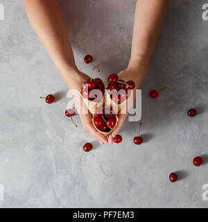 Hände von einer Frau mit knusprigen Waffelbecher mit großen reifen Kirschen Beeren in Ihnen auf einem grauen Tabelle, Platz für Text. Ansicht von oben. Sommer Konzept der Homemad Stockfoto