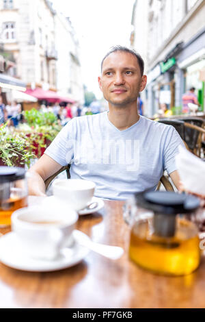 Junge glücklich Mann in European Outdoor Cafe Restaurant sitzen, Leute beobachten Tee trinken mit Teekanne aus Glas, Tassen auf romantischen Datum im Sommer in Stockfoto