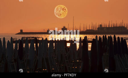 Schöne volle Roter Mond über dem Hafen vom Strand mit Sonnenschirmen gesehen Stockfoto