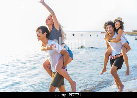 Eine Gruppe von Freunden zu Fuß am Strand entlang, mit Männer, piggyback Ride zu Freundinnen. Glückliche junge Freunde genießen Sie einen Tag am Strand Stockfoto