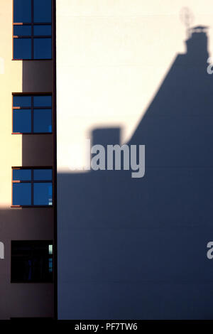 Ein Schatten von einem benachbarten Haus mit Rohren in der Altstadt fällt auf eine glatte Wand mit Fenstern an einem sonnigen Tag Stockfoto