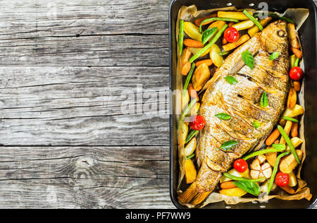 Ganze Fische, die in einer Auflaufform gebacken mit Kartoffeln, Tomaten, grüne Bohnen, Zitrone, Karotten und Basilikum auf einem alten Tabelle, Ansicht von oben, horizontal Stockfoto