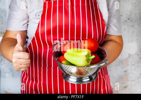 Frau in einem roten Schürze mit einem Sieb mit Gemüse für Salat zeigt Hand Zeichen 'ok'. Das Kochen. Stockfoto