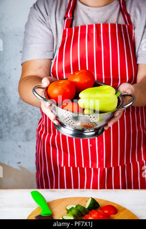 Frau in einem roten Schürze mit einem Sieb mit Gemüse für Salat. Das Kochen. Stockfoto