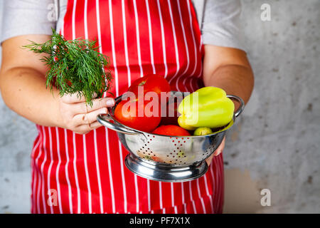 Frau in einem roten Schürze mit einem Sieb mit Gemüse für Salat. Das Kochen. Stockfoto