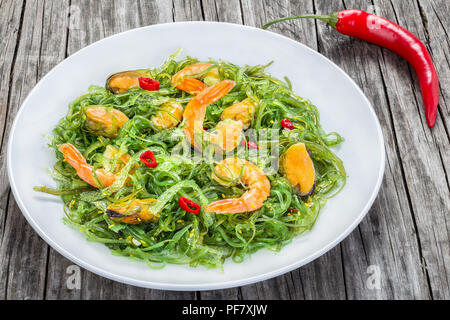 Lecker Salat mit Garnelen, Muscheln, Algen, Chili und Sesam Dressing mit Olivenöl extra vergine auf dem weißen Teller auf die alten hölzernen Tisch, Nahaufnahme Stockfoto