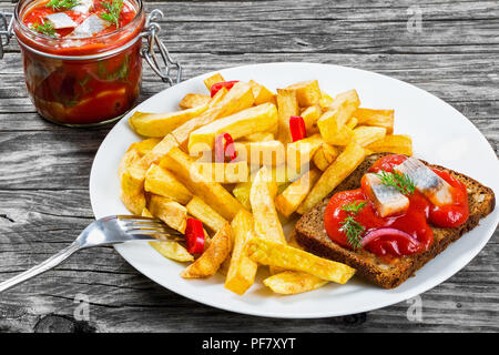 Kartoffel Pommes mit Stücken von Chili auf einem weißen Teller und Scheiben von mariniertem norwegischen Hering in Tomatensoße auf die Stücke von Roggen Brot, ein Glas Stockfoto